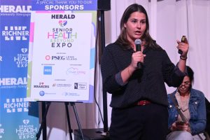 Woman in a dark polka-dot blouse speaks into a handheld microphone beside a sponsorship board at a Senior Health & Beyond Expo event.