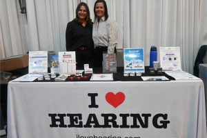 Two women stand behind a booth display with a tablecloth that reads 'I ♥ HEARING' and 'ilovehearing.com' on the front banner, smiling at the camera.