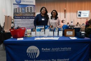 Two women stand behind a Parker Jewish Institute information table at a health expo, with brochures and a large Parker banner in the background.