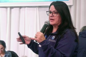 Woman with glasses speaks into a handheld microphone while holding a smartphone during a talk, wearing a purple polo with a logo on it.