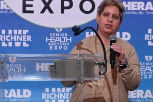 Woman speaks at a clear podium with a microphone during a Senior Health & Beyond Expo event, backdrop includes the expo logo and branding.
