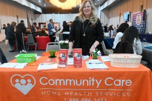 A woman stands behind an orange Community Care booth table at a conference, smiling at the camera with brochures and a basket on the table behind her in a busy expo hall.
