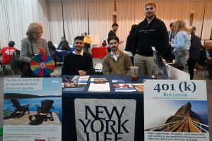 Two men sit at a New York Life information booth with a colorful prize wheel on the left and brochures on the table in a busy event space.