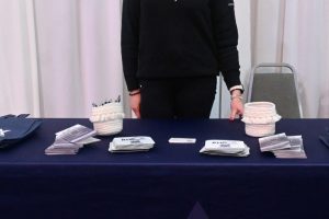 Woman standing behind a dark blue tablecloth branded with 'JMC' at a memorial chapel booth, smiling at the camera with brochures and pens on the table.