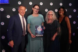 Four adults in formal attire pose together on a black backdrop with circular event logos; the central woman in a teal dress holds a 'WINNER' certificate while the others smile at the camera.