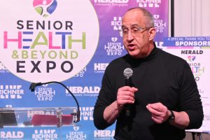 Man in a black turtleneck speaks into a handheld microphone at a Senior Health & Beyond Expo backdrop showing the event logo and sponsor banners