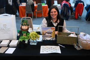 Smiling woman sits behind a table at a booth with a plush leprechaun, a small plant, brochures, and baskets on a black tablecloth.