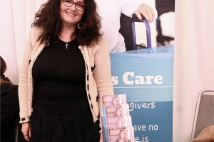 Smiling woman with red glasses standing at a health care booth, with a caregiver banner in the background and brochures on the table.