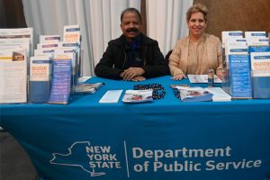 Two staffers sit behind a blue table at a NYS Department of Public Service information booth with pamphlets and pens on display.
