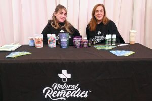 Two women sit behind a table displaying Natural Remedies products and brochures at a booth.