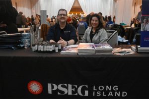 Man and woman sit behind a table with stacks of brochures and bottled products at a PSEG Long Island booth in a busy event hall.