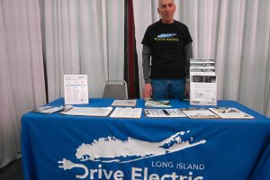 Man in a black Drive Electric shirt stands behind a blue-clothed information table at an EV booth with brochures and a tall display next to him.
