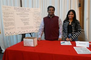 Two smiling presenters stand at a red-table booth with a large poster about dementia awareness titled 'Remember Me' behind them.