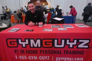 Smiling man sits behind a red GymGuyz table at a trade show, brochures and a mug visible on the table with attendees in the background.