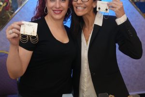 Two women at a jewelry booth smiling, each holding pair of large hoop earrings on display cards.
