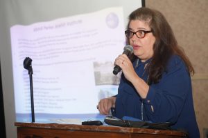 Woman with red glasses speaking into a handheld microphone at a podium, presenting a projected slide behind her.