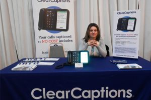 Woman sits at a ClearCaptions promotional booth with banners, brochures, and a captioned phone on the table.