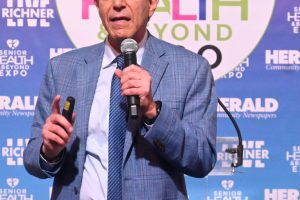Man in a blue blazer speaks into a handheld microphone on a stage with a colorful health expo backdrop behind him.