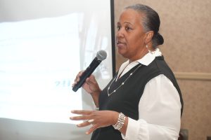 African American woman in a white blouse and black vest speaking into a handheld microphone during a presentation.