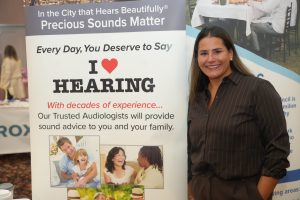 Smiling woman standing beside a large banner about hearing care, which features the 'I ❤️ HEARING' slogan and related text.
