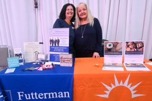 Two smiling women stand behind a booth with blue and orange tablecloths, displaying law firm brochures and posters labeled Futterman Lanza LLP.