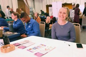 Smiling blonde woman in a gray top sits at a table with brochures and a pink-phone on the table; event booths and other attendees are in the background.
