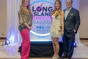 Three people pose for a photo at a long island awards event in front of a white draped backdrop with the Herald logo