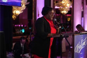 African American woman in a red dress and black blazer speaks at a podium with a microphone at an awards event stage.