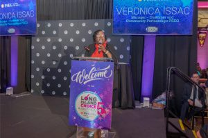 Black woman in a red blouse speaks at a podium on stage during the Long Island Choice Awards ceremony, with blue sponsor banners behind her.