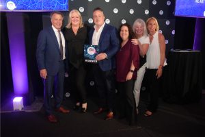 Group of six smiling adults at an awards event, with a man in a blue suit holding a plaque that says WINNER; backdrop features logo badges and blue stage lighting.
