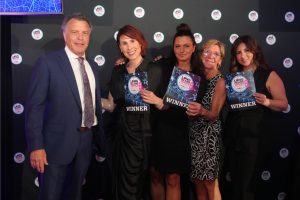 Five people pose with winner signs at an awards event, standing in front of a logo backdrop. (man in blue suit on the left, four women in black attire)
