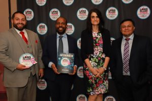 Group of four professionals in formal attire posing in front of a Long Island Choice Awards backdrop; the second man holds a winner plaque/brochure.