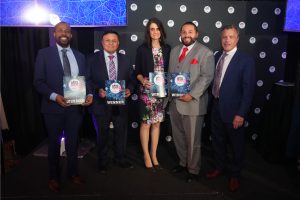 Five adults in suits pose on stage holding WINNER certificates at an awards event.