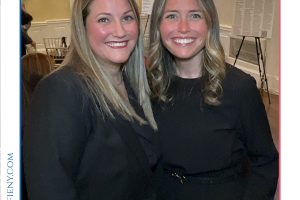 Two smiling women in black outfits posing together at a formal event, with event branding and logo visible around the frame.