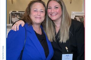Two women posing together at a professional event, one in a royal blue blazer and the other in black, smiling for the photo with a name badge visible on the right.