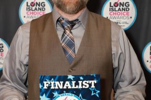 Smiling man in a brown vest and tie holding a finalist certificate at the Long Island Choice Awards backdrop.