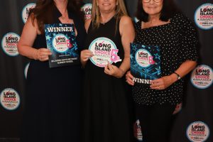 Three women posing with 'Long Island Choice Awards' winners, holding booklets, in front of a branded backdrop.