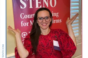 Smiling woman in a red patterned dress with glasses poses with arms raised in front of a red 'STENO' banner at a legal awards event (Top Lawyers of Long Island).