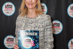 Woman in a patterned dress smiling while holding a 'WINNER' award sign at the Long Island Choice Awards backdrop.