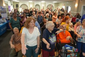 Crowd of seniors at an indoor event dancing and cheering, some raising arms, with tables and banners in the background.