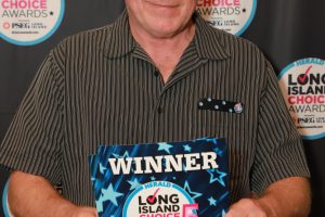 Man in a striped shirt smiling while holding a 'WINNER' award card for East Meadow Upholsterers at the Long Island Choice Awards, with event logos in the backdrop.