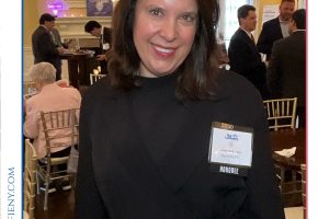 Smiling woman at a formal awards event, wearing a dark top and a badge reading HONOREE, with other guests in the background at a decorated venue.