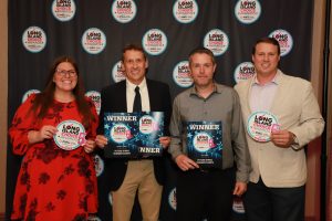 Four people pose at a Long Island Choice Awards backdrop; two men in the center hold 'WINNER' plaques while others hold round award disks.
