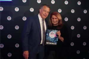 Man in a blue suit and a woman with red glasses posing, holding a 'WINNER' award plaque at an event backdrop.