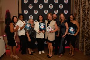 Group of nine people posing together at an awards event, holding WINNER plaques with a branded backdrop behind them.