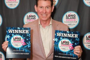 Man in a beige blazer smiles, holding two Long Island Choice Awards winner certificates against a backdrop of circular award logos.