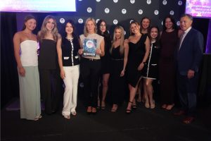 Group of eleven people on a stage at an awards event; the central woman holds a WINNER certificate, with a backdrop of logos and lights.