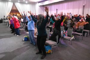 Group of adults standing and raising arms in a large hall during a synchronized exercise session; chairs and bags nearby.