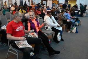 Crowd of people seated in a conference hall waiting area, many holding papers and bags in their laps and at their feet.