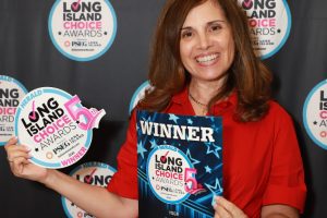 Smiling woman in a red dress holds a 'WINNER' award poster and a circular 'Long Island Choice Awards' sign, standing in front of a branded backdrop.